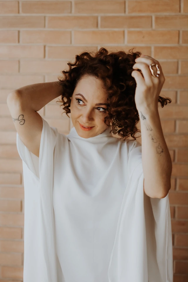 Ingrid com as mãos junto aos cabelos, diante de uma parede de tijolos